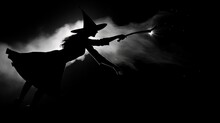 Woman Dressed As A Witch Flying In The Black Night Charming With A Wand With White Sparkle On The Background Of Clouds With Moonlight In Silhouette Style