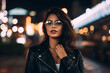Young trendy dressed hipster in eyewear with night city light reflection fascinated with beautiful illumination, gorgeous woman in leather jacket wondering while standing on urban background
