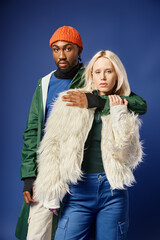 african american man embracing blonde woman in stylish winter attire on blue backdrop, multiethnic