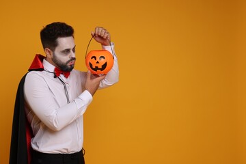 Poster - Man in scary vampire costume with fangs and pumpkin bucket on orange background, space for text. Halloween celebration