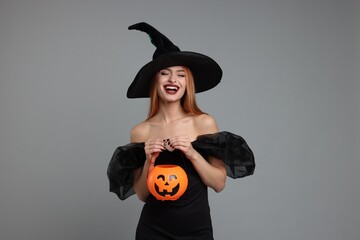 Wall Mural - Happy young woman in scary witch costume with pumpkin bucket on light grey background. Halloween celebration