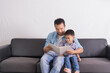 © janonkas - happy asian Father and son reading book together while sitting on couch at home. happy family, father day