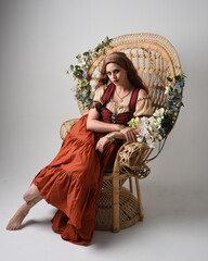 Sticker - Full length portrait of beautiful red haired woman wearing a medieval maiden, fortune teller costume.  Sitting pose, with gestural hands reaching out. isolated on studio background.