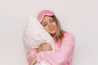 © Марина Демешко - A young sleepy blonde woman in a pink pajamas and sleep mask with her eyes closed hugs a pillow isolated on a white background. Insomnia, sweet dreams, cuddling with a pillow