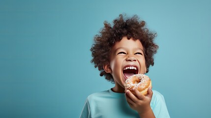 A little boy with a smile is eating a donut on a blue background wall. The child is having a good time with the donut. It's a fun time to have sweet food at home.