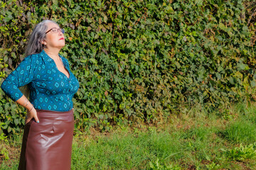 Wall Mural - Elegant older adult woman standing in garden with her eyes closed and head tilted back sunbathing, brown skirt, blue blouse, long gray hair, hands on her waist, calm expression on her face