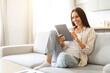 © Prostock-studio - Smiling beautiful woman relaxing with digital tablet on couch at home