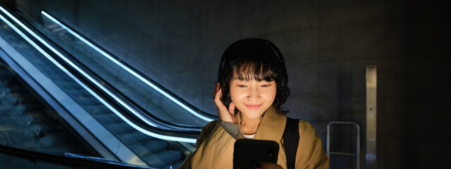 Wall Mural - Portrait of beautiful korean girl in headphones, stands near escalator, travels, commutes home on public transport, holds smartphone, poses with mobile phone