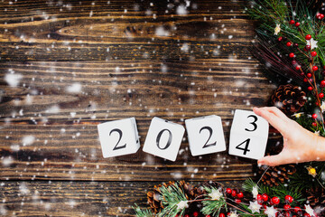 Woman's hand flipping New Year's 2023 wood calendar blocks to 2024. Christmas tree lights, pine branches, red winter berries and snow. Top view.