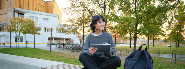 Wall Mural - Young girl, korean artist or art student sits in park with digital tablet, draws with graphic pen, scatches a design or project, looks around