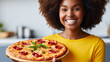 A young, smiling African-American woman is holding a large, freshly baked pizza in a kitchen setting. She appears to be happy and excited about the pizza