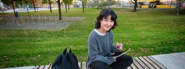 Wall Mural - Young asian girl with graphic pencil and tablet, sits in park on bench, draws scatches, does her homework outdoors