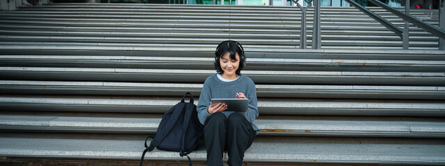 Wall Mural - Young smiling digital artists works on her project using tablet and graphic pen, listens music in headphones and sits on stairs in public space, works on project