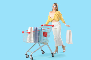 Poster - Stylish happy young woman with shopping bags and cart on blue background