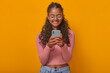 Young optimistic pretty Indian woman teenager holding phone in hands and typing message in college group chat enjoying sharing funny pictures in mobile messenger stands posing in orange studio.