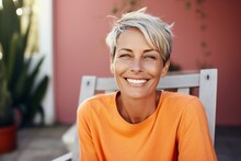 Smiling Middle-aged Woman Sitting On The Terrace Of Her Cozy Country House. Attractive Blonde Female With Short Hairstyle Enjoys Silence Of Her Beautiful Garden. Adult Lady With Happy Smile Relaxed.