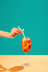 Female hand putting straw into Aperol spritz cocktail isolated over green background. Refreshment, aperitif. Concept of alcohol drinks, party, holidays, bar, mix. Poster. Copy space for ad