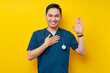 Smiling professional young Asian male doctor or nurse wearing a blue uniform and stethoscope waving hand to say hi, hello, greeting patient, hand on chest isolated on yellow background