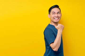 Wall Mural - Smiling professional young Asian male doctor or nurse wearing a blue uniform standing confident while pointing a finger at copy space isolated on yellow background. Healthcare medicine concept