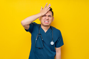 Wall Mural - Mistake professional young Asian male doctor or nurse wearing a blue uniform looking at camera and hand on his head isolated on yellow background. Healthcare medicine concept