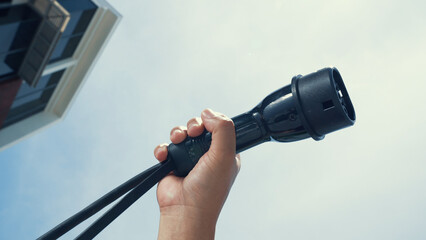 Closeup hand grasping an EV plug for electric vehicle with the midday sky in the background as progressive idea of alternative sustainable clean and green energy for environmental concern.