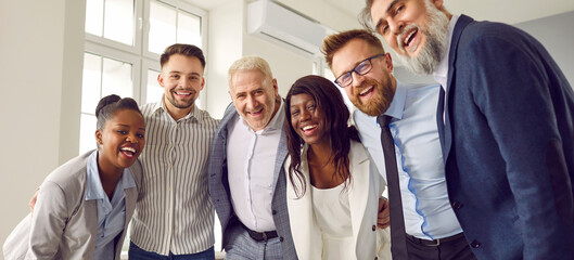 Group of business multiethnic people men and women looking at camera hugging and smiling indoors. Company employees team or group of staff standing in the office confidently and cheerfully. Banner.