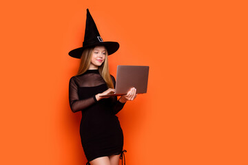 Photo portrait of lovely blonde teen lady hold laptop typing message dressed black halloween garment isolated on orange color background