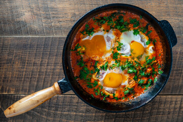 Wall Mural - Delicious shakshuka with eggs, tomato and parsley in a cast-iron pan, closeup, top view. Shakshuka - traditional israeli tomato stew with eggs