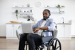 Young person in casual wear carrying portable computer while sitting in wheelchair in studio apartment. Close up of positive african american adult looking at camera.