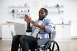 Young african american person in casual wear with portable computer while sitting in wheelchair in studio apartment. Positive adult man having video call, while sitting at kitchen.