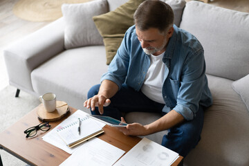 Poster - Cheerful senior man in casual clothing using digital tablet while sitting on the sofa at home.