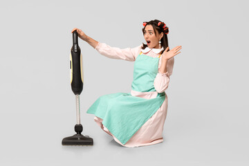 Poster - Portrait of shocked young housewife in apron with vacuum cleaner on grey background