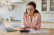 © Prostock-studio - Focused woman with laptop, cellphone and calculator in kitchen
