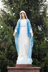 Statue of Virgin Mary. Mother Mary outdoor. Open arms love. Religious background. Catholic religion holy person. Blessed Mother Mary with pine tree behind. Coniferous background.