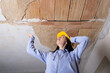 A young woman wearing a hard hat gazes up at a damaged hole in the ceiling, where the plaster has crumbled due to moisture damage. She is engaged in the restoration, repair old apartment