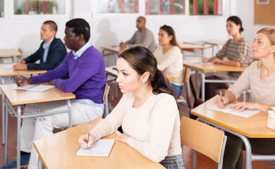 Canvas Print - Focused young attractive brunette listening to lecture in classroom with group of adult people. Postgraduate education concept