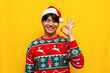 © Богдан Маліцький - young asian man in christmas sweater and santa hat smiling and showing ok gesture on yellow isolated background