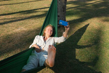 A Young Girl With Curly Blond Hair Lies In A Hammock In Nature, Takes A Selfie On A Smartphone And Enjoys Life, With Her Bare Foot In The Foreground.