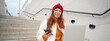 Smiling redhead woman with mobile phone and laptop, sitting on stairs outside building, connects to public wifi, using smartphone and computer