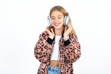 Pleased Beautifu blonde kid girl enjoys listening pleasant melody keeps hands on stereo headphones closes eyes. Spending free time with music