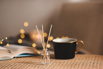 Wall Mural - Home liquid fragrance in glass bottle with cup of coffee and paper book on rattan table over xmas lights close up. Winter holiday season. Aromatherapy.