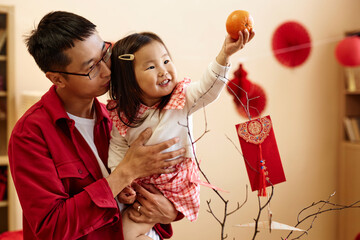 Wall Mural - Waist up portrait of happy Asian girl holding tangerine while celebrating Chinese New year with father at home, copy space Have overflowing abundance every year