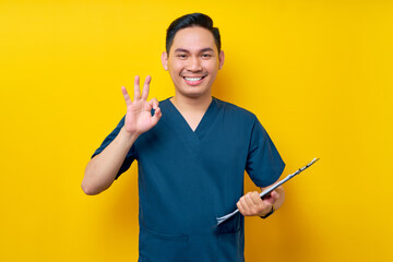 Wall Mural - Smiling professional young Asian male doctor or nurse wearing a blue uniform holding clipboard and showing okay sign isolated on yellow background. Healthcare medicine concept