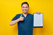 © Bangun Stock Photo - Smiling professional young Asian male doctor or nurse wearing a blue uniform holding clipboard with blank paper and showing thumbs up or like isolated on yellow background. Healthcare medicine concept