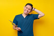 Stressed professional young Asian male doctor or nurse wearing a blue uniform holding clipboard and scratching his head with hand isolated on yellow background. Healthcare medicine concept