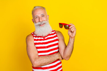 Poster - Photo portrait of old man take off brand new sunglass in striped red overall smiling at summer season isolated on yellow color background