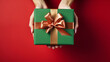© Malambo/Peopleimages - AI - Hand holding a green gift box with gold bow, on a red table. Valentine, Christmas or birthday