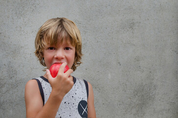 Adorable caucasian blond little 6 years old boy biting happy a red apple with vitamins looking to the camera outside isolated concrete background. Healthy eating in childhood concept. Copy space