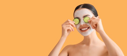 Poster - Beautiful young woman with cucumber slices and facial mask on orange background with space for text. Cosmetology concept