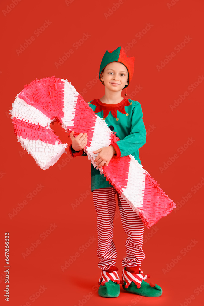 Cute little boy in elf costume with candy cane pinata on red background